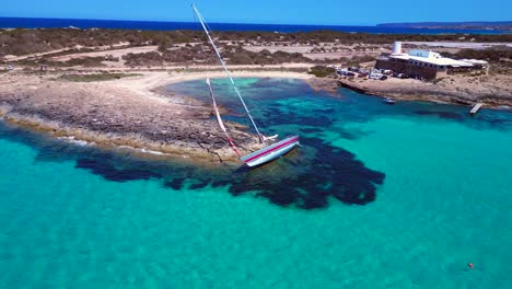 sailboat stranded on cliff near the coast of formentera island, spain, during a summer sunny day. smooth aerial view flight descending drone