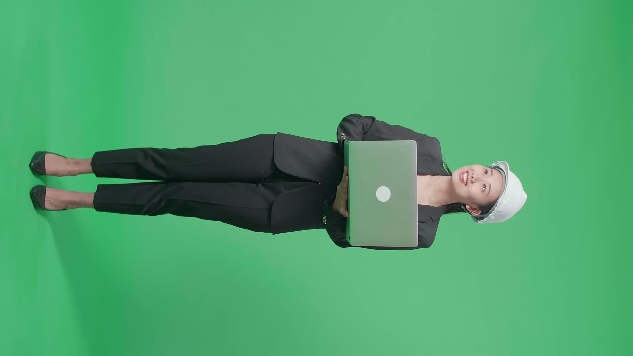 Full Body Fo Smiling Asian Female Engineer Looking Around And Shaking Her Head While Using Laptop In The Green Screen Studio