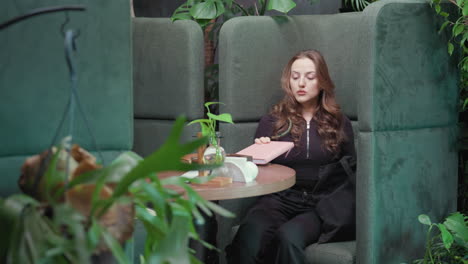 blonde woman dressed in black sits on green cushioned chair inside plant-filled interior, reaching into black bag to pull out books as she settles into calm cozy booth with soft light