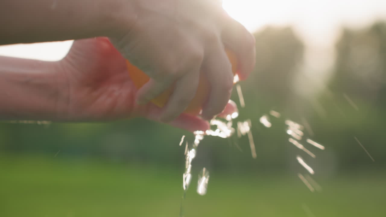 close up of kid hands washing orange under running water in park with blurred trees background and sun flare, healthy summer snack preparation, playful outdoor moment, fresh fruit cleaning and care