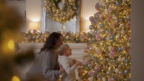 little boy decorate christmas tree with balls and ornaments, mother lifting him up