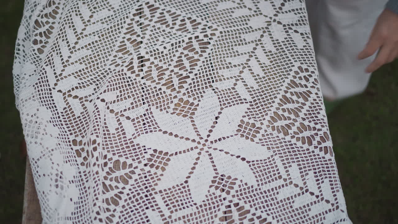 Kaukasiske hænder placerer en blondedug, et vintage hæklet dækkeservietmønster afsløres, husmor glatter stoffet ud over en vejrbidt træbænk, havegræs i baggrunden, nærbillede af stingdetaljer.