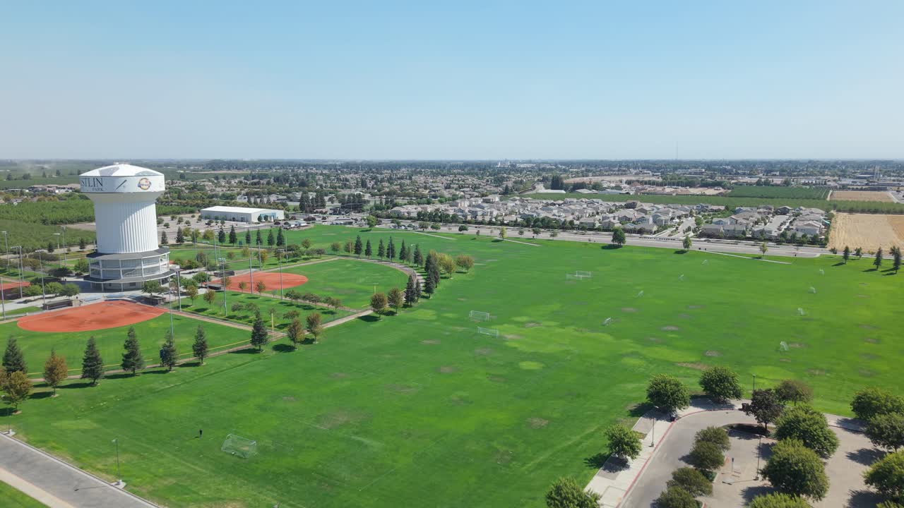 Tracking aerial shot passing water tower, green fields, trees, and residential area