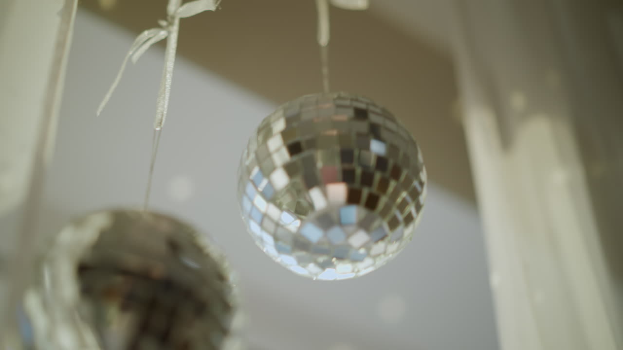 Close up shiny silver disco balls hanging indoors reflecting sunlight on surrounding walls, glittering mirrored surface creating sparkling light patterns, decorative festive atmosphere in modern home interior