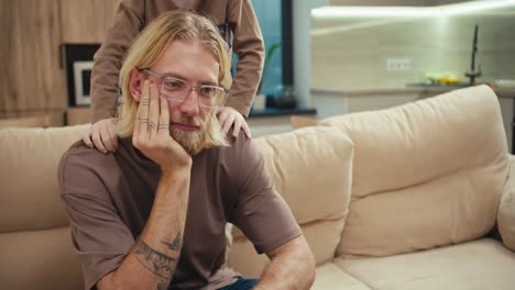 A-tortured-blond-man-with-a-beard-and-glasses-sits-on-the-sofa-while-his-little-albino-son-pesters-him-and-wants-to-play-with-the-man-at-home-in-a-modern-apartment.-Tired-man-with-glasses-cannot-pay-enough-attention-to-his-little-albino-son-in-a-modern-apartment