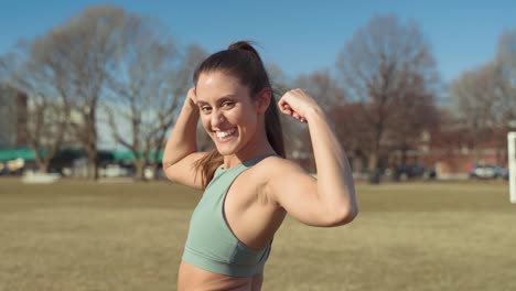 joven y bella mujer fitness flexiona los músculos del brazo mientras se gira para sonreír a la cámara sintiéndose orgullosa de su fuerza, poder y confianza después del entrenamiento en el parque