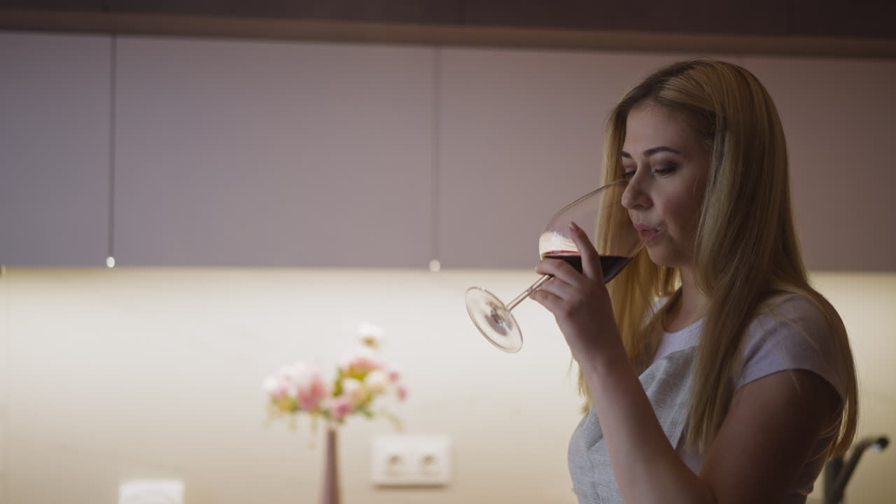 Young woman drinks wine and smiles cooking in kitchen