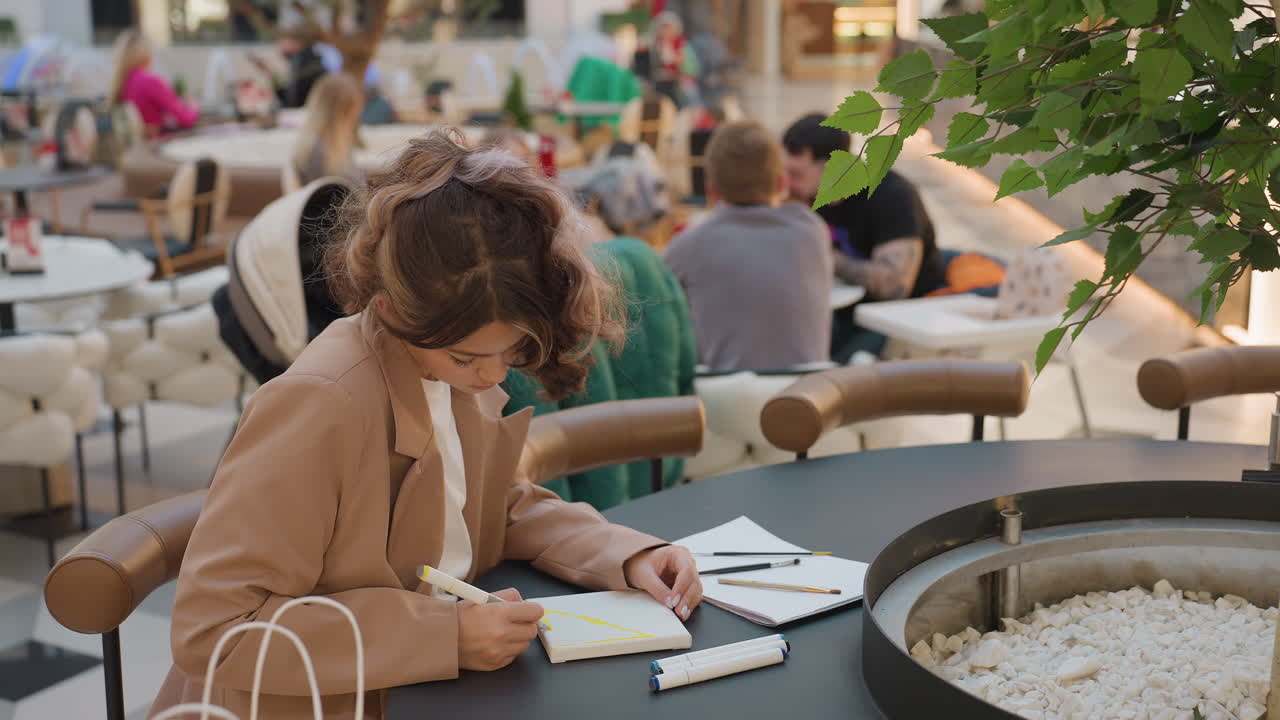 붐비는 인파 속에서 계획을 세우는 여성, 도시의 번잡한 쇼핑객들에게 둘러싸여 일정을 준비하는 여성, 활기찬 도시 환경 속 쇼핑몰 벤치에서 플래너에 집중하는 젊은 여성