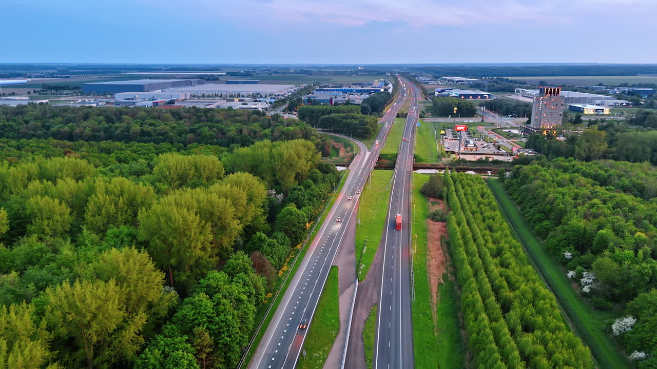 Green fields by highway, NL. An aerial view captures a highway surrounded by lush greenery and industrial areas in the Netherlands during sunset
