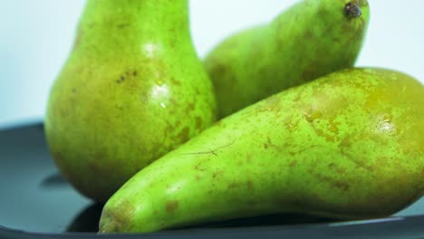 peras verdes grandes y frescas giran lentamente sobre un plato negro sobre fondo azul claro, concepto de comida saludable, primer plano extremo, cámara gira hacia la izquierda