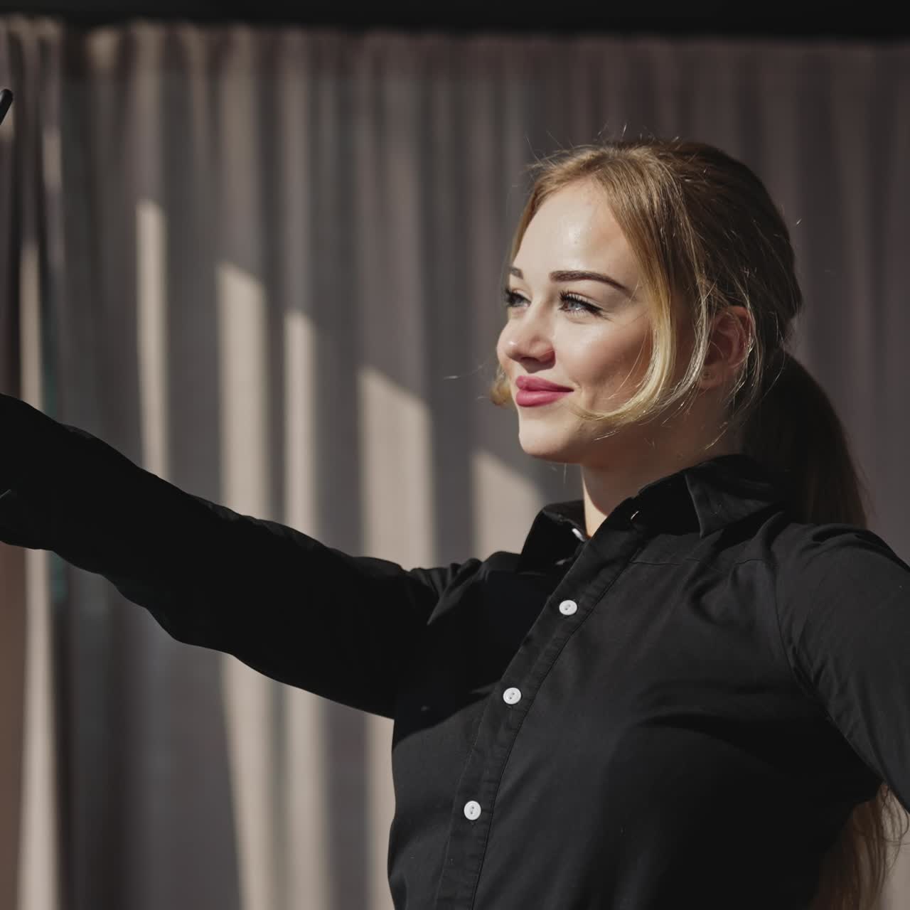 Young woman taking selfie in front of the window. Long-haired woman looks at her phone and closes her eyes from the bright sun shining