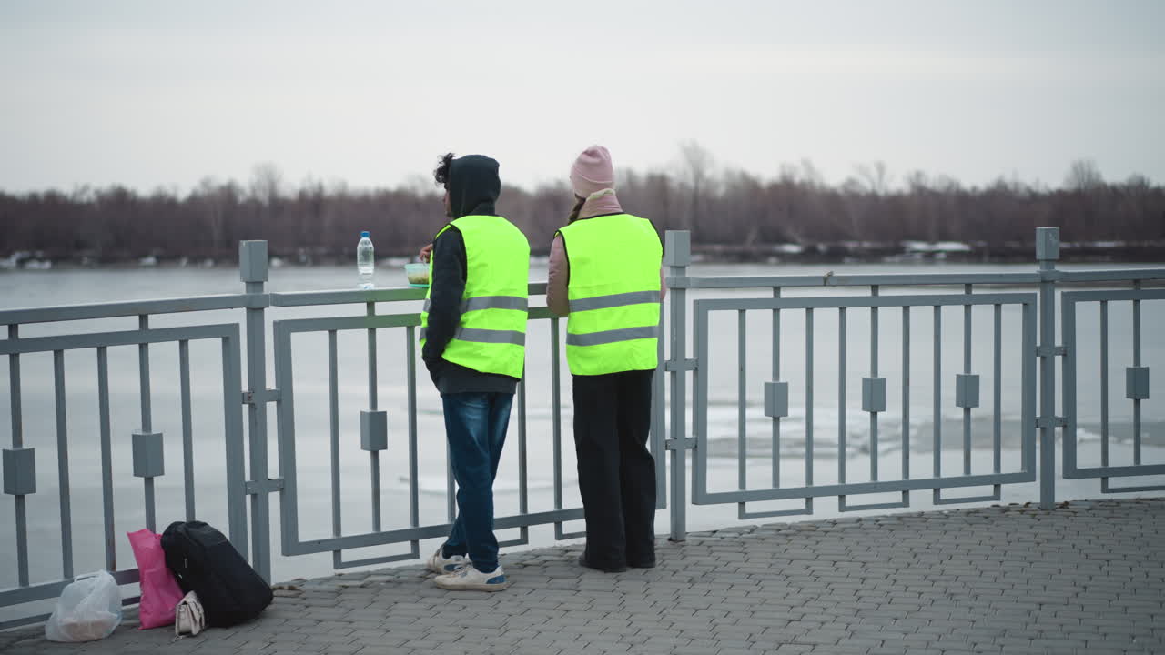 Two people in reflective safety vests stand at riverside railing eating food from containers in cold weather, with bags and backpack on ground, overcast sky, and bare trees along frozen water background