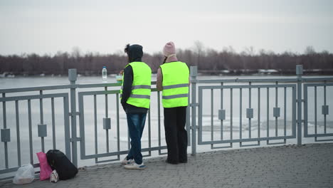 two people in reflective safety vests stand at riverside railing eating food from containers in cold weather, with bags and backpack on ground, overcast sky, and bare trees along frozen water background