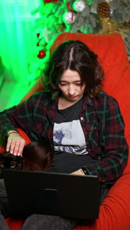girl in checkered shirt sits in the chair with a cat and laptop. lady working remote online at christmas. vertical video.