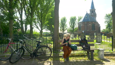 a female cyclist resting on a bench drinking some water in front of a church in friesland, zurich