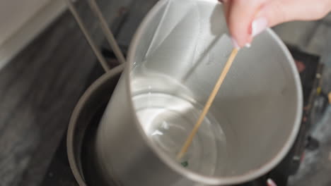close up of metal container placed inside steaming pot with stick stirring clear greenish liquid as part of preparation process on rustic stove in kitchen, focused view of melting or mixing operation