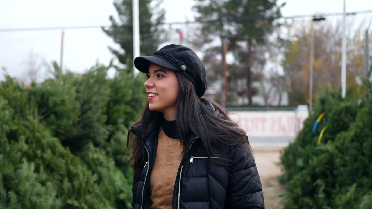 una mujer joven comprando una decoración de árbol de navidad de vacaciones de temporada en un lote con muchas especies de árboles festivos