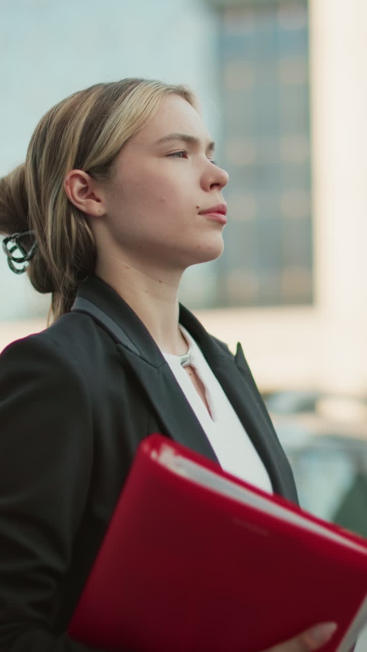 Side view of remote worker with hair clip holding red folder and coffee cup glancing left with parked cars and modern office building background during workday in bright urban setting