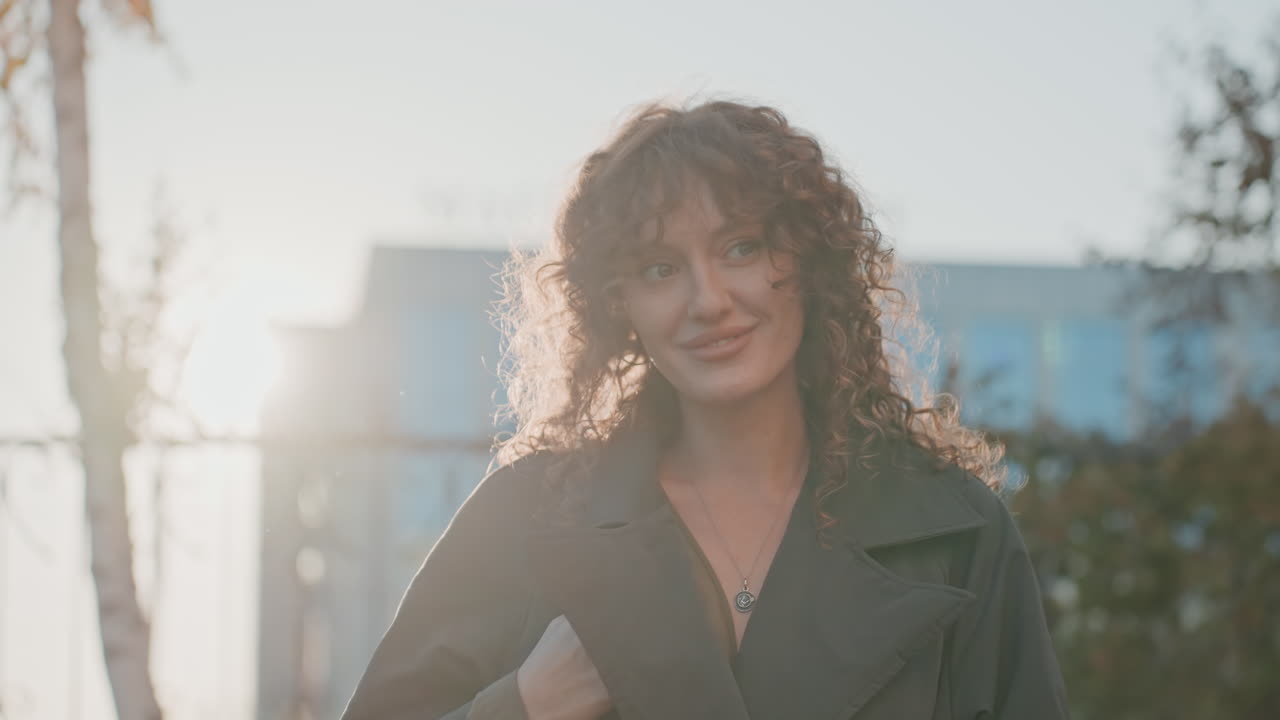 Retrato urbano ensolarado de mulher com cabelo cacheado, casaco trench coat e cerca de ferro. Folhas de outono, contraluz quente e um brilho cinematográfico suave criam um clima contemplativo. O enquadramento próximo captura um sorriso sutil.