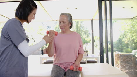lifting dumbbells, senior asian woman receiving assistance from physiotherapist during home therapy