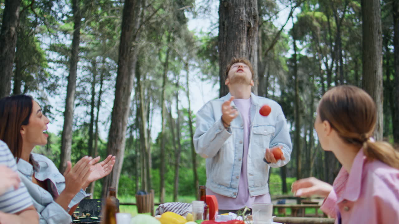 Friends having fun forest picnic. Playful man juggling with vegetables in park