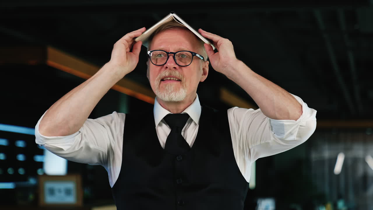 Old man wearing white shirt and black vest holds his paper notebook on his head. Positive smiling man in glasses joking, fooling around. Blurred backdrop.