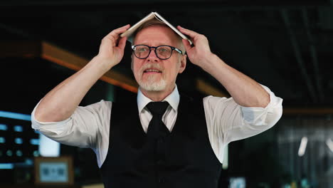 old man wearing white shirt and black vest holds his paper notebook on his head. positive smiling man in glasses joking, fooling around. blurred backdrop.