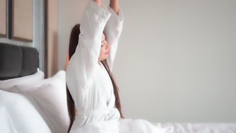 attractive woman in a terrycloth bathrobe stretches sitting in a huge hotel suite bed