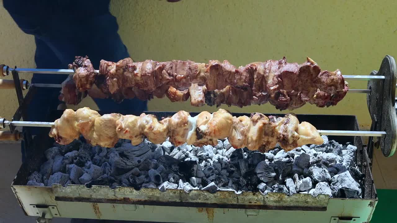 Primo piano di uno spiedino da BBQ riposto su calore intenso su una griglia a carbone. Carne che cuoce lentamente con fiamme visibili, fumo e una consistenza sfrigolante. Ideale per immagini di street food, cucina mediterranea.