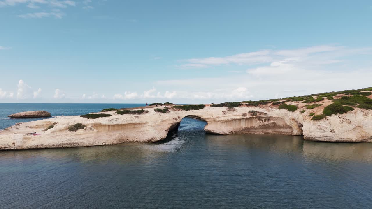 Aerial view of S'Archittu's natural rock arch in Sardinia, Italy. Waves crash against the beach, showcasing a stunning coastal landscape. Perfect for travel, nature, and scenic beach footage.