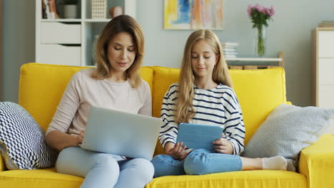 blonde mother and daughter sitting on yellow couch while they talking and using laptop computer and tablet in the living room 2