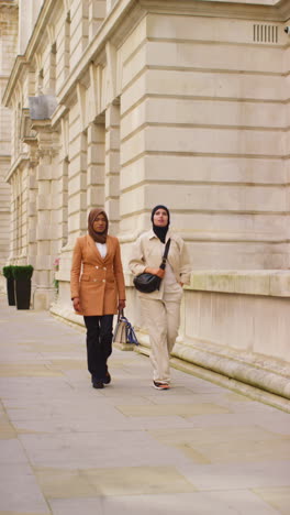 Vertikales-Video-Von-Zwei-Muslimischen-Geschäftsfrauen-Mit-Hijabs-Und-Modernen-Geschäftsanzügen,-Die-Auf-Dem-Weg-Zur-Arbeit-An-Städtischen-Bürogebäuden-Vorbei-Gehen-1