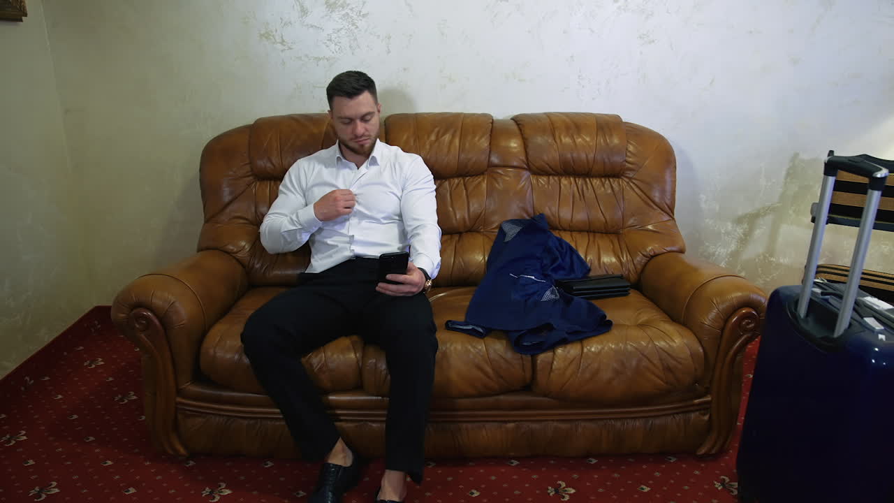 Tired businessman is resting in hotel room. Young man is sitting on sofa and looks at mobile. Comfortable trip away from home.