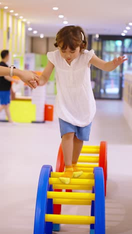 a mother holds her 5-year-old daughter's hand for help and support as the little girl carefully balances and walks across a colorful ladder obstacle, developing motor skills