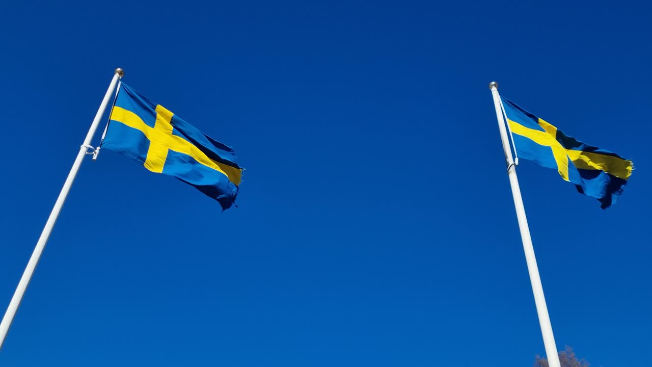 Two Swedish flags whipping vigorously in strong wind against a clear blue sky, representing national pride, heritage, and Scandinavian identity