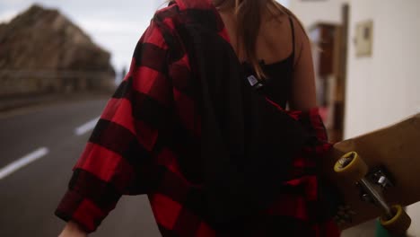 cropped footage woman in plaid coat on a cloudy day at walking, holding a skateboard, slow motion. unrecognizable girl walks by asphalt road