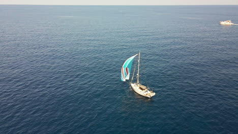 Velero-Con-Velas-De-Colores-Navegando-En-El-Mar-Azul-Durante-La-Puesta-De-Sol-Del-Verano-En-Cerdeña,-Italia---Toma-Amplia-De-Drones-Aéreos