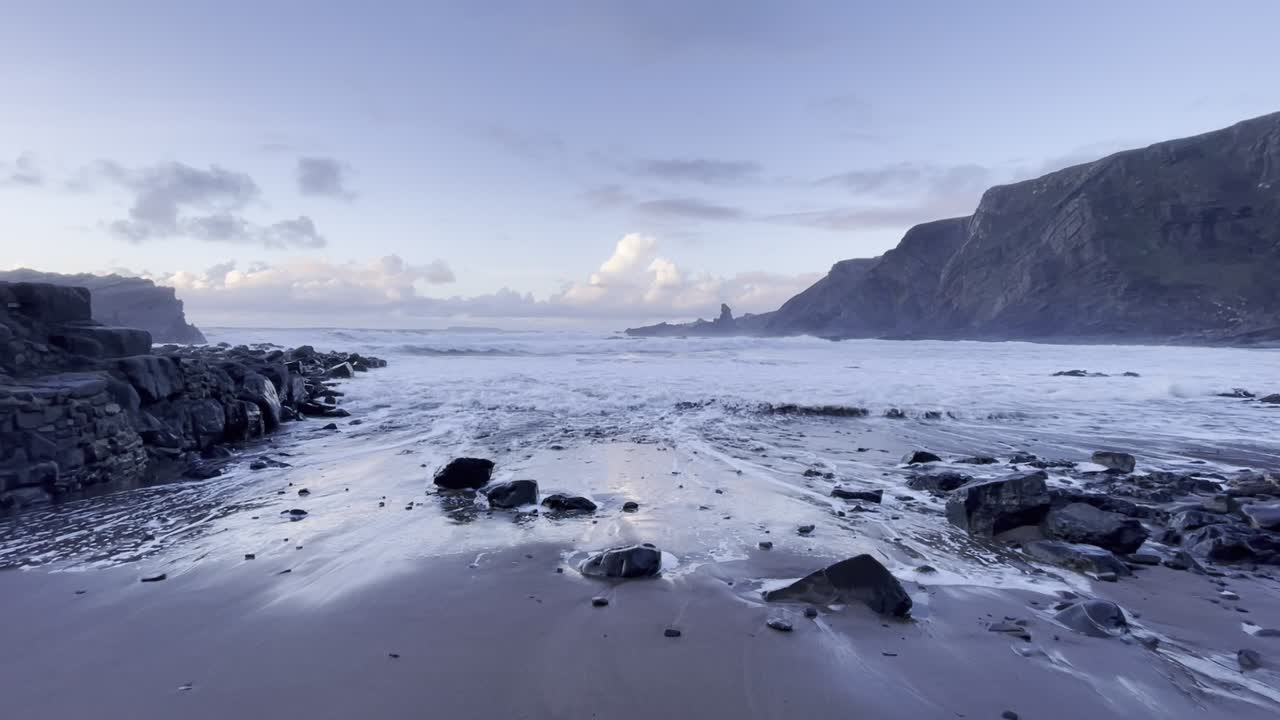 Aerial footage captures the rugged coastline of Cornwall with waves crashing onto a rocky beach. The serene atmosphere is enhanced by soft lighting, highlighting the dramatic cliffs and views