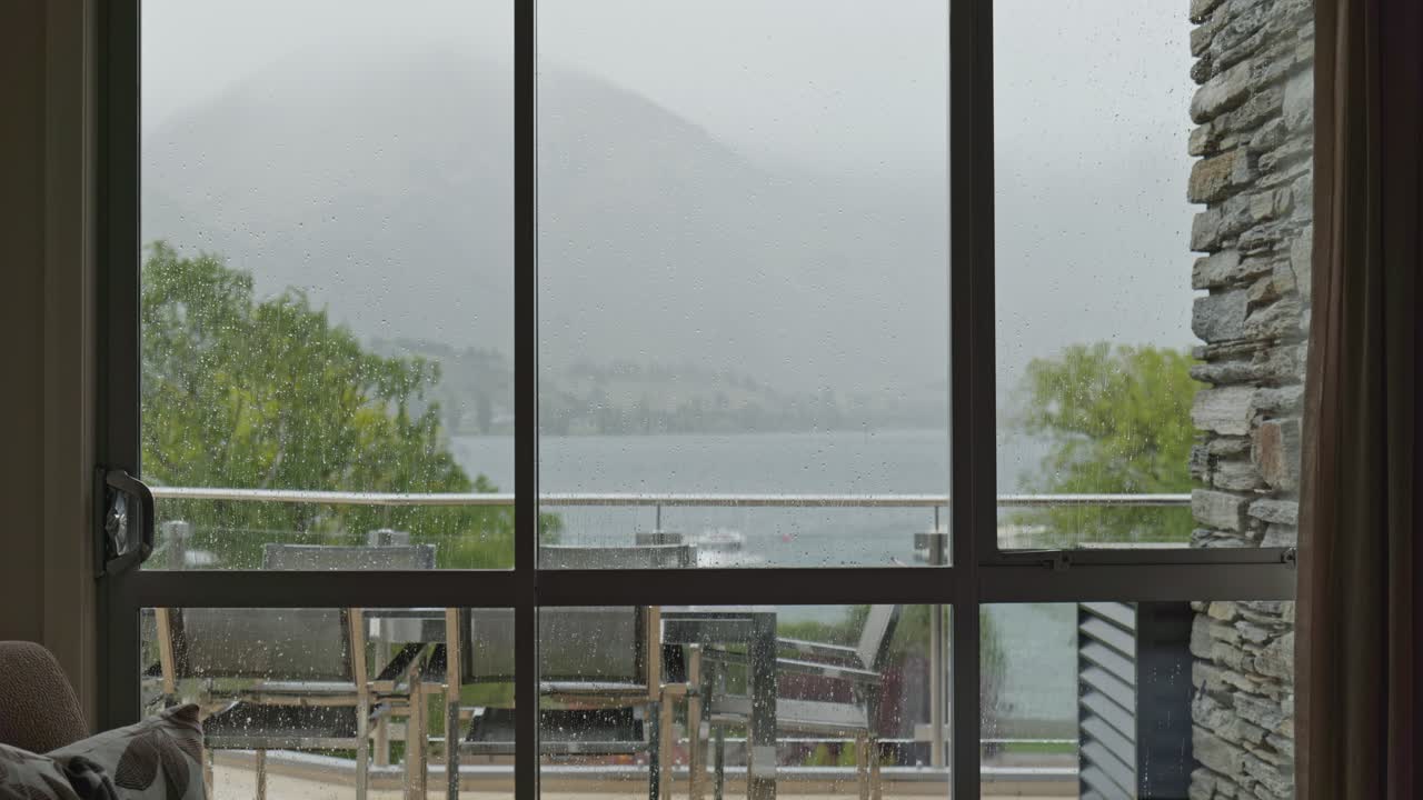 View from a Wanaka hotel room during rain, showing balcony chairs and an approaching boat on the lake through a wet window