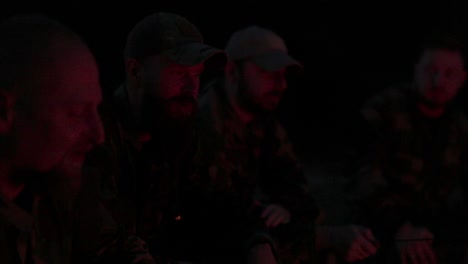 a team of men in camouflage clothing is sitting in the field at night, warming themselves by a lit campfire, frying sausages for dinner, a manly outing, relaxing after work, service, hunting