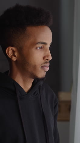 thoughtful african-american man in hoodie looks out of window standing in spacious room close side view. person suffers from loneliness at home