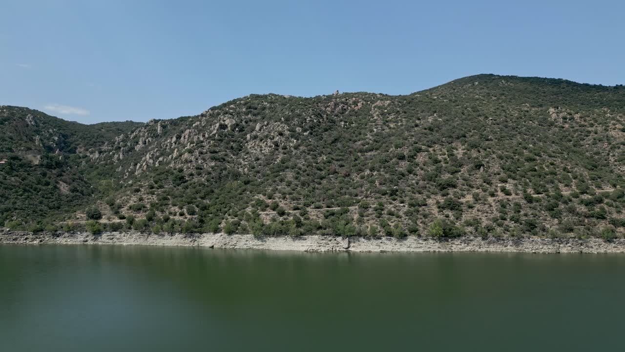 Explore the serene coastline and lush hills around the tranquil lake at midday in this picturesque natural setting. Lac Vinça, France