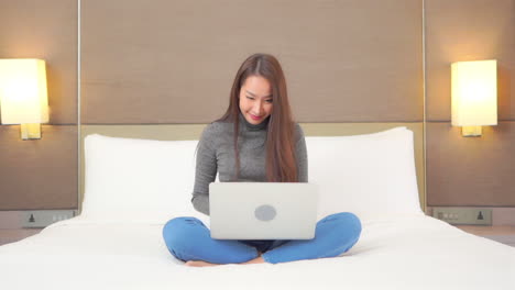 a young woman comfortably works on her laptop while sitting on a huge hotel suite bed