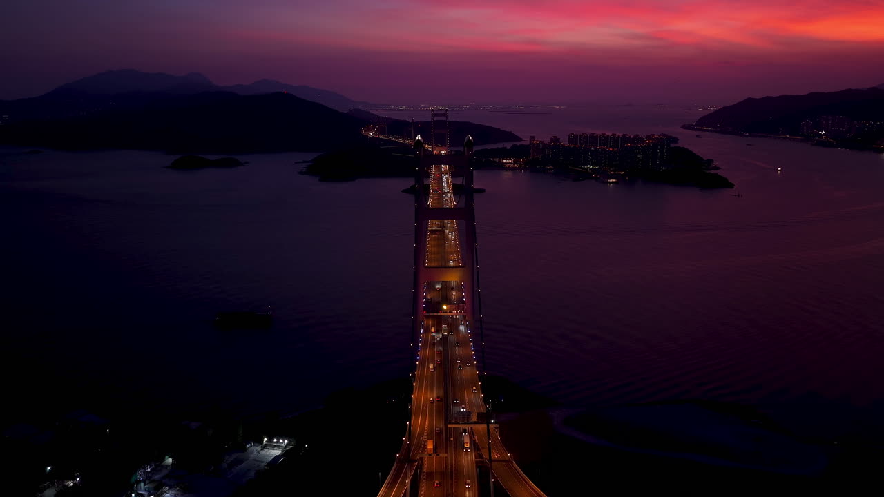 Cinematic Aerial view of Tsing Ma Bridge in Hong Kong at sunset, spanning over calm waters with city skyline, mountains, and ships in the distance, creating a serene urban and natural landscape scene