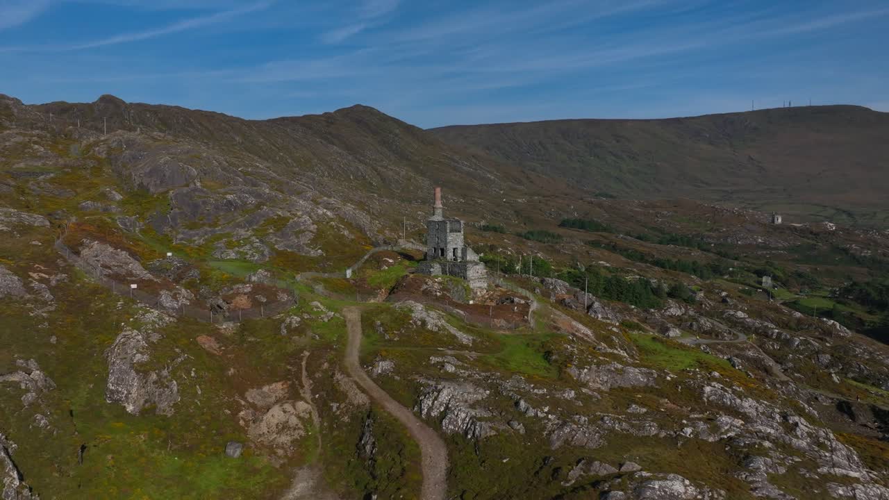 Copper Mine, Allihies, County Cork, Ireland, September 2024. Drone pushes in and orbits clockwise from a wide view of mountain slope towards the historic ruined Engine House under a bright blue sky.
