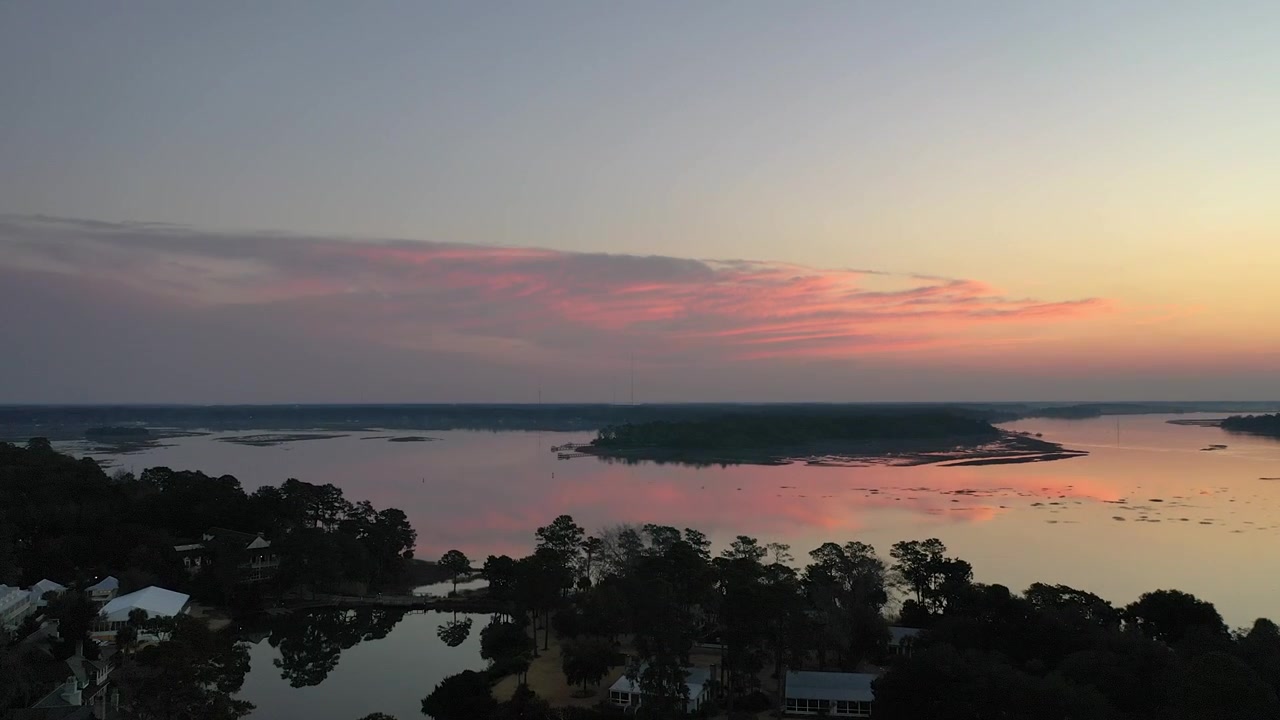 Imágenes de dron sobrevolando un paisaje fluvial tranquilo al atardecer, revelando exuberantes bosques verdes y casas a lo largo de la orilla con hermosas nubes rosadas reflejándose en el agua en calma.