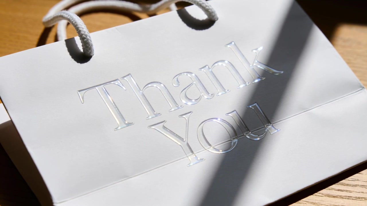 Sacchetto di carta bianco su tavolo di legno, con la luce solare che si sposta, proiettando un'ombra sulla scritta "Thank You" in rilievo e sui manici.