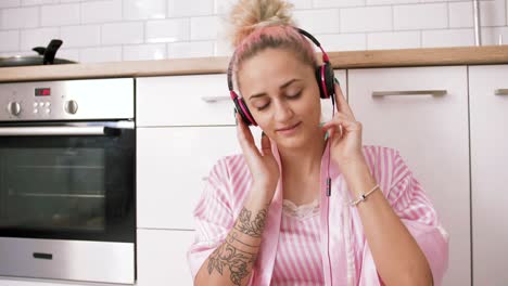 happy young woman with pink hair sitting on the floor on kitchen wearing pink pajamas and listening to music with headphones