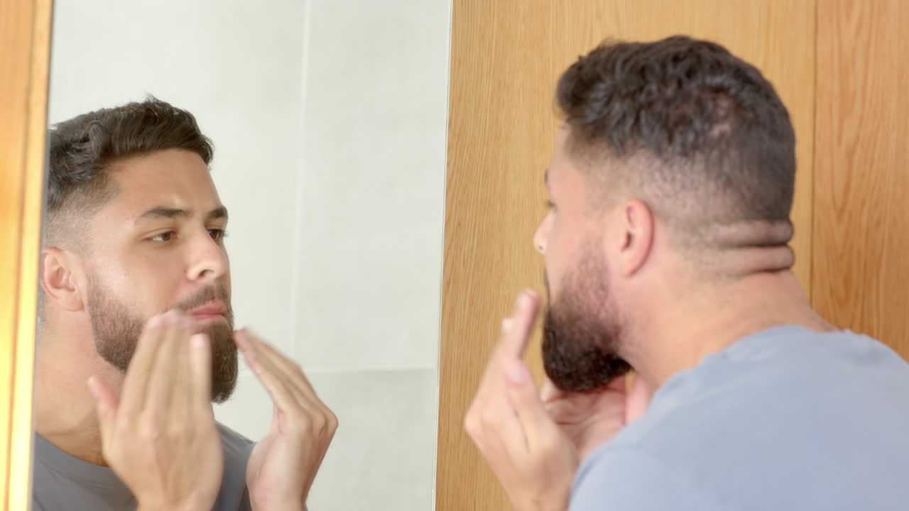Man looking in mirror, touching face and examining skin in bathroom