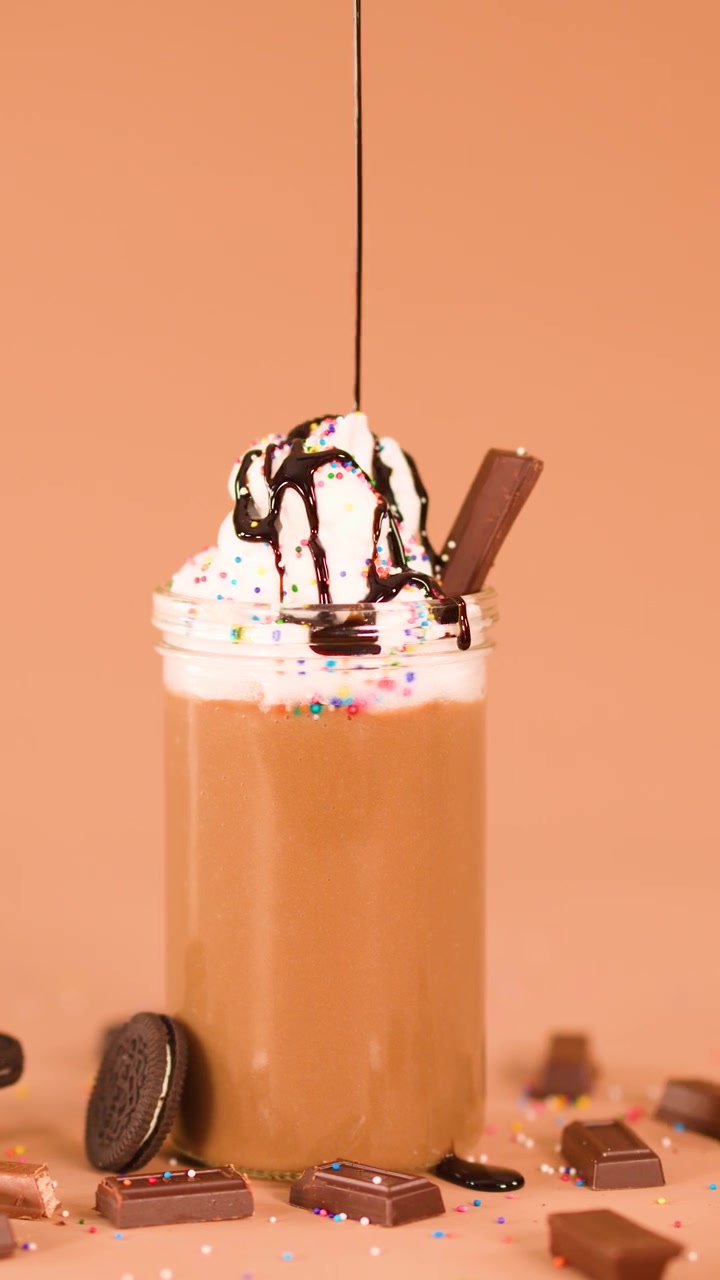Sciroppo al cioccolato che cola su un milkshake con panna montata, biscotti, codette di zucchero e cioccolato, su sfondo marrone